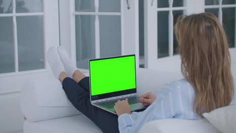 Eine-Junge-Frau,-Die-Auf-Der-Couch-Liegt,-Blickt-Auf-Den-Bildschirm-Des-Laptops-Mit-Grünem-Bildschirm-Und-Nickt-Mit-Dem-Kopf.-Einen-Videoanruf-Tätigen